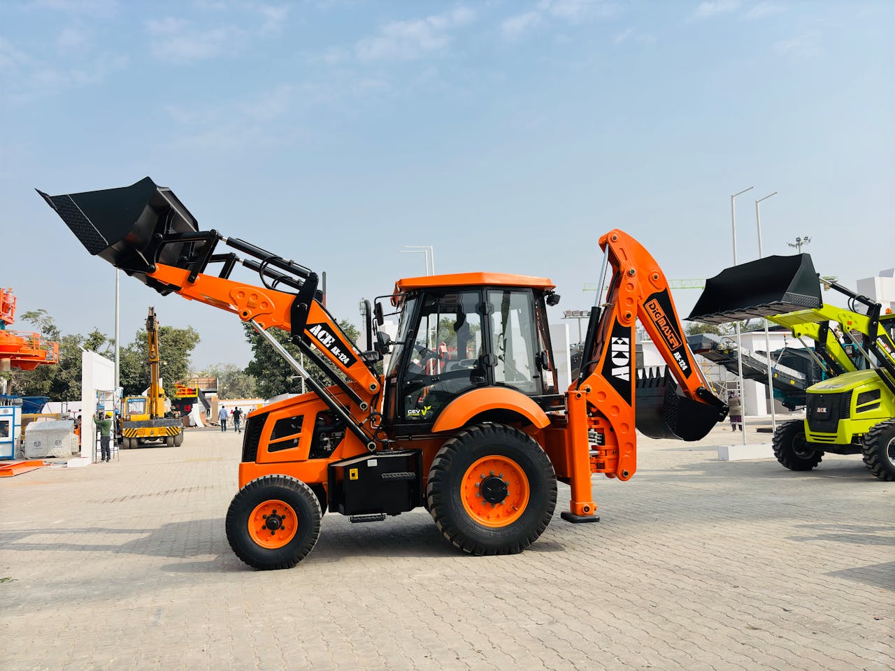 Bright orange ACE AX 124 backhoe loader on display at an outdoor exhibition in India.